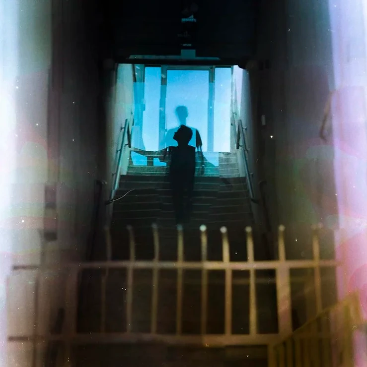 A silhouetted figure at the top of a stairwell washed in blue and violet light.